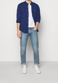 Tmavě modrá košile na knoflíky přes bílé tričko, spojená se světle modrými zvednutými džíny a bílými teniskami, stojící na neutrálním pozadí.