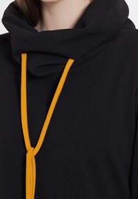 Svart hoodies med gul snörning. Har ett mjukt, texturerat tyg och en lös passform med en justerbar halsringning.