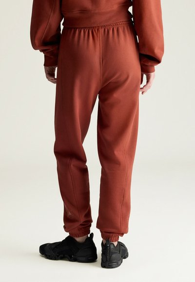 Osoba nosząca joggery w kolorze rdzy z elastycznym stanem i mankietami, w połączeniu z czarnymi butami sportowymi, stojąca na jasnym tle.