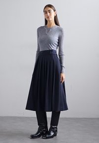 Top gri cu maneci lungi din tricot, cu decolteu tip henley, purtat cu o fustă plisată bleumarin. Botine negre cu cataramă completează vestimentația.
