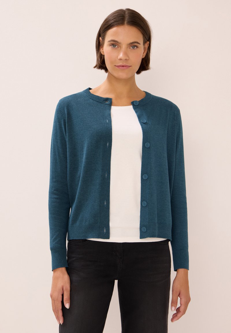 Een turkooise cardigan van zachte stof, met een ronde halslijn, knoopsluiting en lange mouwen, gecombineerd met een wit shirt eronder.