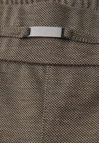 Bruine textuurstof met een ruitpatroon, voorzien van een metalen sluiting en stikdetails bij de taille.