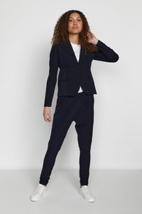 Navy blue blazer with structured shoulders and lapels, paired with matching tapered jogger pants. White t-shirt underneath. White sneakers.