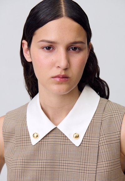 Gros plan sur un jeune avec des cheveux noirs raides, portant une robe à motifs avec un col blanc et des boutons dorés, affichant une expression neutre.