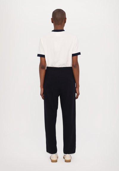 White polo shirt with navy collar and cuffs paired with black trousers. Trousers have a relaxed fit, featuring a decorative white trim at the hem.