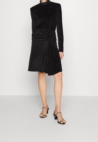 Robe en velours noir avec des manches longues, un col montant et un design froncé à la taille. Présente des accents scintillants et une jupe évasée. Sandales à talons noires.