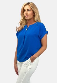 Niebieska bluzka z krótkim rękawem, z detalem guzikowym i luźnym krojem. Połączona z białymi spodniami. Gładka tekstura, casualowy fason.