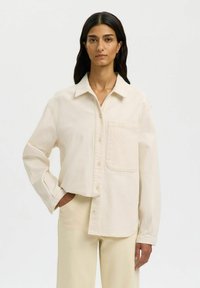 Femme aux longs cheveux bruns portant une chemise oversize crème boutonnée et un pantalon assorti, debout avec une main dans la poche.