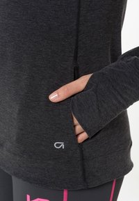 Dunkelgraues, langärmliges Sportoberteil mit einer Seitentasche, Daumenlöchern und einem kleinen Logo am Saum. Weicher, dehnbarer Stoff mit minimalen Nähten.