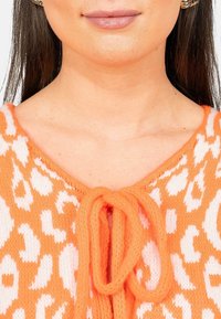 Maglione lavorato a maglia arancione con stampa leopardata bianca, caratterizzato da un scollo a V e una chiusura con fiocco frontale. La texture è morbida e lavorata in modo rilassato.