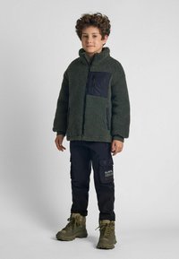 Veste en polaire verte avec une poche noire, portée avec un pantalon cargo noir et des bottes olives. La veste a un col montant et une coupe décontractée.