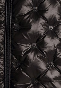 Chaqueta de abrigo negra con una textura brillante y acolchada. Presenta símbolos de paz bordados y un cierre frontal con cremallera.