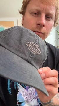 Pelēks kokvilnas beisbola cepure ar noapaļotu kroni un stingru malu, ar baltu izšūtu logotipu priekšpusē. Mīksta tekstūra, ikdienišķs stils.