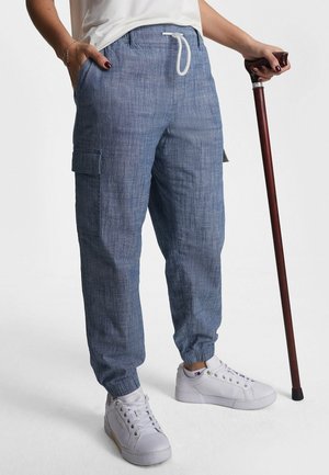 Personne portant un pantalon cargo bleu à cordon de serrage et des baskets blanches, tenant une canne en bois à poignée courbée dans la main droite.