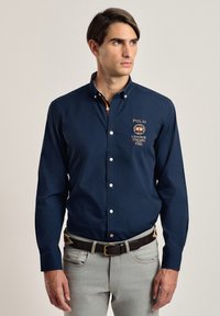 Camisa botón azul marino con logo bordado, mangas largas y cuello estructurado. Combinada con jeans gris claro y un cinturón marrón.