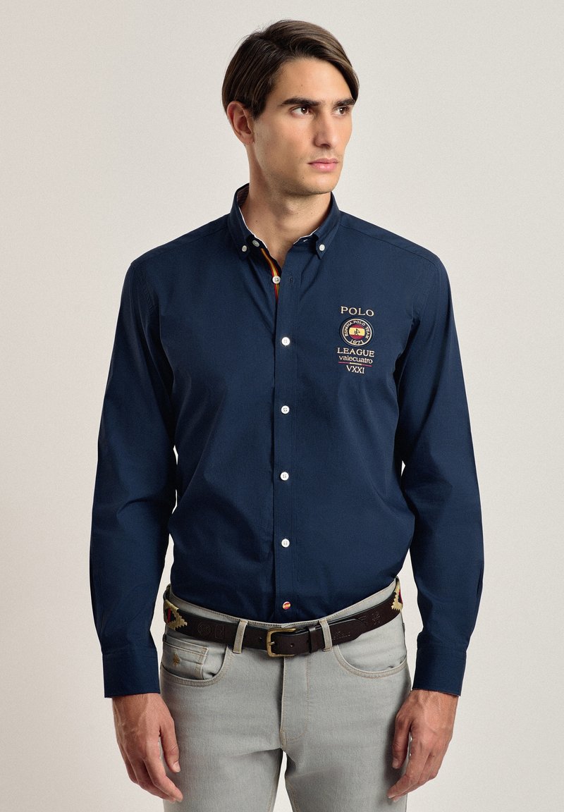 Camisa botón azul marino con logo bordado, mangas largas y cuello estructurado. Combinada con jeans gris claro y un cinturón marrón.