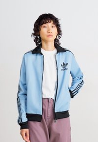 Asmuo vilkintis šviesiai mėlyną Adidas sportinį švarką su juodomis juostelėmis, baltą marškinėlį ir migdolų spalvos kelnes, stovintis prieš paprastą baltą foną.