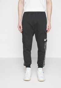 Mustat cargo-housut, joissa on sivutaskut, valkoiset raidat ja Nike-logo reidessä, yhdistettynä valkoisiin lenkkareihin.