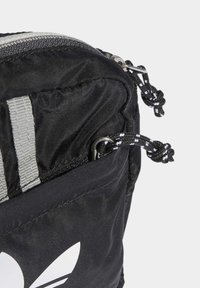 Borsa in tessuto nera con logo Adidas bianco, dotata di tasca con zip e cordini per la zip in bianco e nero con estremità a nodo.