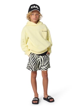 Sweat à capuche jaune avec poche avant, short à motif zèbre noir et blanc, et sandales noires avec le logo "KARL" sur la lanière.