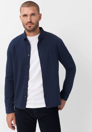 Mann mit kurzen Haaren und Bart, der ein dunkelblaues Hemd über einem weißen T-Shirt und schwarzen Hosen trägt, steht vor einem einfarbigen hellen Hintergrund.