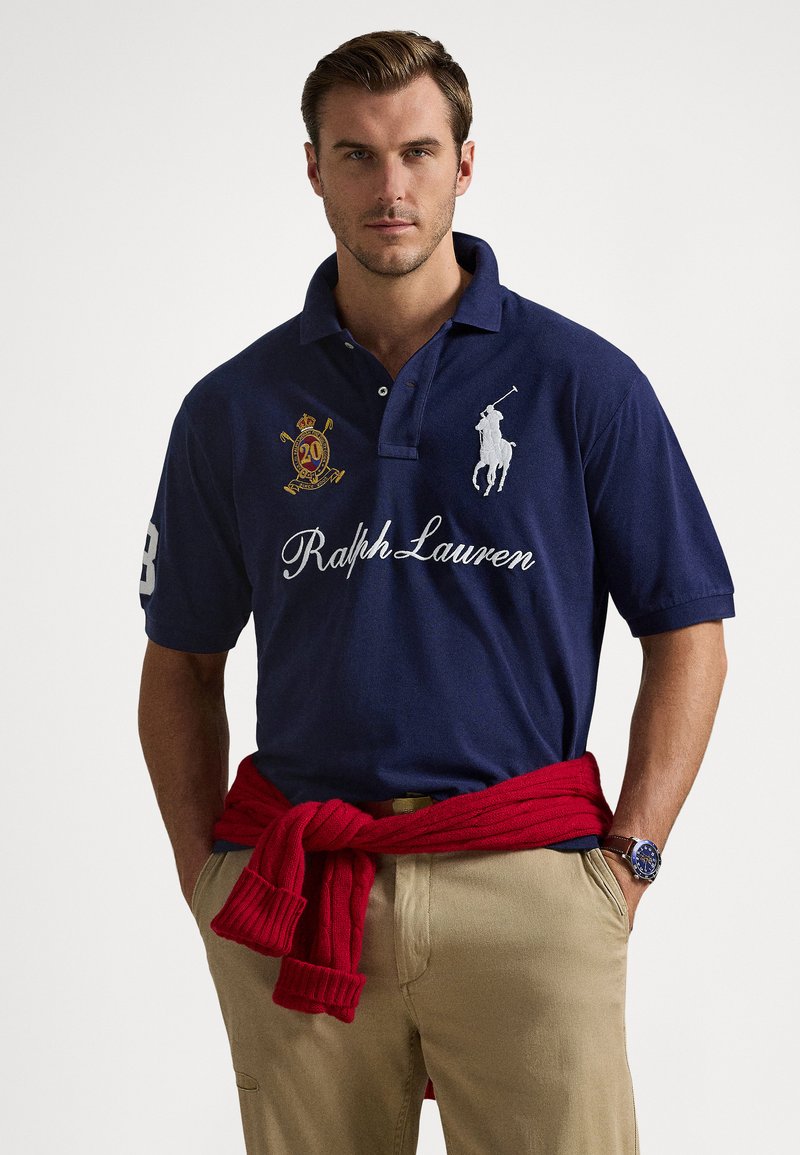 Moški, ki nosi temno modro polo majico Ralph Lauren z vezenimi logotipi, bež hlače, rdeč pulover, privezan okoli pasu, in modro uro na levi zapestju.