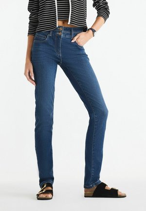 Hubené džíny v tmavě modrém denim s viditelným šitím, dvoubotkovým zapínáním, předními kapsami, doplněné černými plochými sandály se zlatými akcenty.