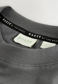Grauer Ausschnitt des Kleidungsstücks mit schwarzem Innentape, das mit "BAKER BY TED BAKER" beschriftet ist, sowie Stofftags, die den Markennamen und "Hergestellt in Bangladesch" anzeigen.