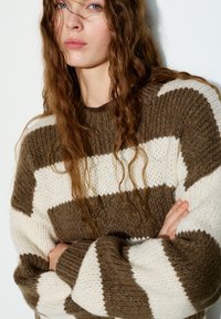 Pull en maille épaisse avec rayures horizontales marron et crème. Présente un col rond et un tissu texturé, avec des manches retroussées.