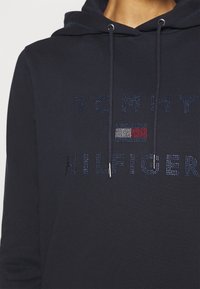 Granatowa bluza z kapturem z logo "TOMMY HILFIGER" zdobionym kryształkami. Miękki materiał, regulowany kaptur ze sznurkami oraz metalowe zakończenia sznurków.