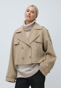 Blond naine, kes kannab üle suuruse beeži kahekordse nööbiga lühikest mantlit beeži kõrge kaelusega sviitri ja mustade pükste peal, seistes heledal taustal.