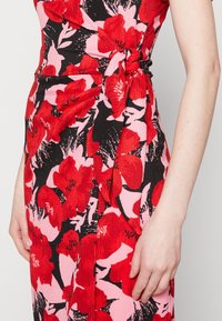 Gros plan d'une robe portefeuille avec de grands motifs floraux rouges et roses sur fond noir, nouée à la taille, montrant le torse et le bras d'une femme.