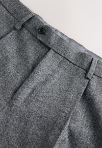 Pantalon en laine grise texturée avec passants de ceinture, plis et fermeture à bouton noir sur un fond clair.