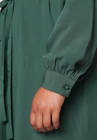 Zelené, lehké šaty z látky s dlouhými, nafouklými rukávy. Detail rukávu s manžetou má zapínání na knoflík. Měkká textura a splývavý design.