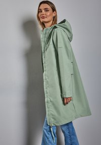 Parka imperméable vert clair avec capuche, boutons-pression, poches latérales et coupe légèrement oversize. Présente un logo sur la manche.