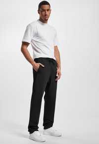 White cotton t-shirt paired with black drawstring pants, featuring side pockets. Model stands against a plain white background. White sneakers on feet.