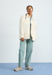 Lys beige skjorte med knapper, blå topp, teal-stripete bukser, grå sneakers. Store speilsolbriller og hoop-øredobber. Glatt, avslappet stil.