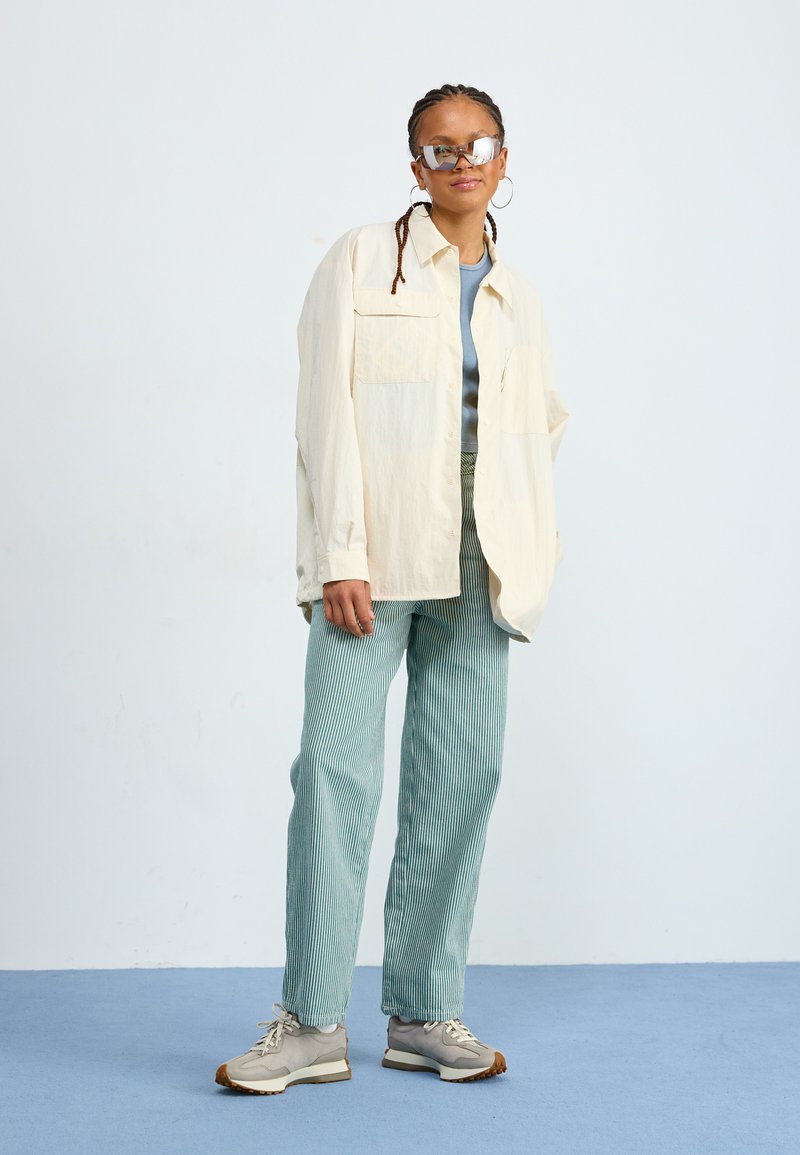 Helles beigefarbenes Hemd mit Knöpfen, blauer Oberteil, türkis gestreifte Hose, graue Turnschuhe. Große Spiegelsonnenbrille und Creolen. Lässiger, eleganter Stil.