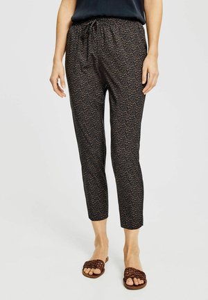 Femme portant un pantalon noir fuselé avec de petites taches marron et des sandales tressées ouvertes marron, debout devant un fond uni.