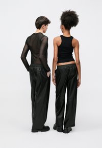 Δύο άτομα στέκονται πλάτη με πλάτη, φορώντας ένα μαύρο διάφανο μακρυμάνικο τοπ και ένα μαύρο αμάνικο τοπ, συνδυασμένα με φαρδιά μαύρα παντελόνια και χοντρά μπότες.