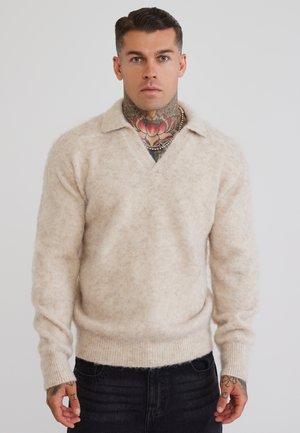 Mand med hals- og håndtatoveringer bærer en beige, blød V-hals trøje og sorte bukser og står mod en ensfarvet lys baggrund.