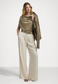 Metallic gold Oberteil mit drapiertem Ausschnitt, hochgeschnittene cremefarbene weite Hose und einem passenden goldenen Gürtel. Hält eine goldene Jacke.