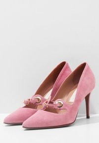 Souliers à talons hauts en daim rose avec des orteils pointus, des côtés découpés et des accents en métal doré à l'avant. Texture lisse et design élégant.