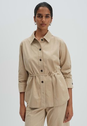Frau trägt beiges, tailliertes Hemd mit Knöpfen und passenden Hosen, steht vor einem schlichten hellgrauen Hintergrund.