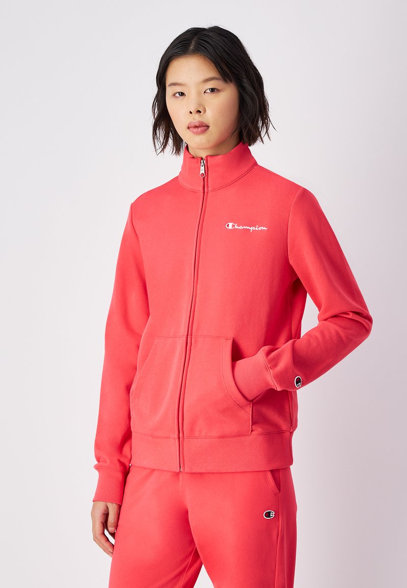 Sudadera roja con cremallera hecha de tejido suave, con bolsillos laterales, puños acanalados y un pequeño logo en el pecho. Combinada con pantalones de chándal a juego.