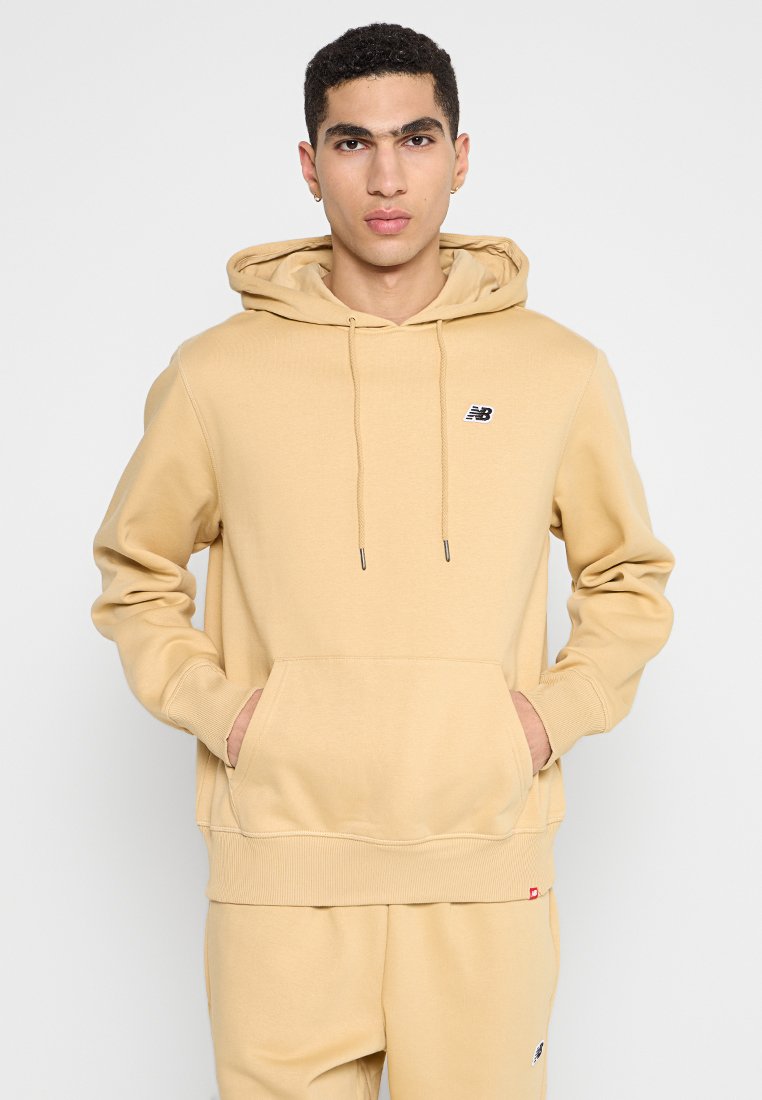 Sweatshirt beige à capuche en tissu doux, avec une poche kangourou, des poignets côtelés et un petit logo noir sur la poitrine.