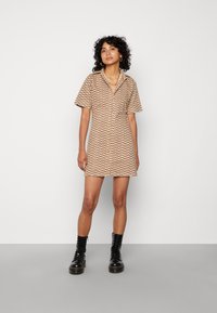 Vestido de manga curta em bege com padrões ondulados em castanho, apresentando uma gola e fecho de botão, usado com botas de combate pretas.