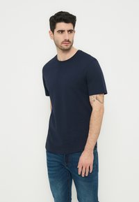 Άνδρας με σκούρα μαλλιά, φορώντας μπλε κοντομάνικο μπλουζάκι και μπλε τζιν, στέκεται μπροστά σε λευκό φόντο, κοιτάζοντας στο πλάι.