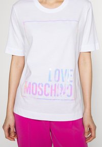Bílý bavlněný tričko s krátkými rukávy, s holografickým potiskem "LOVE MOSCHINO" na spodní části, lemovaným barevným obdélníkovým rámečkem.