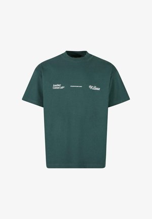 Zaļš kokvilnas T-krekls ar apaļu kakla izgriezumu. Uz tā ir balti iespiesti teksti: "Vēl viens Kokvilnas Laboratorija," "Greznība ir jauna greznība" un "Neskaidra sezona."